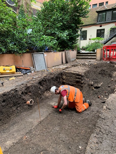 Photograph of a man in orange high visibility protective clothing and a hard hat, kneeling to uncover the next layer in an exposed trench in the garden of Stationers' Hall. 